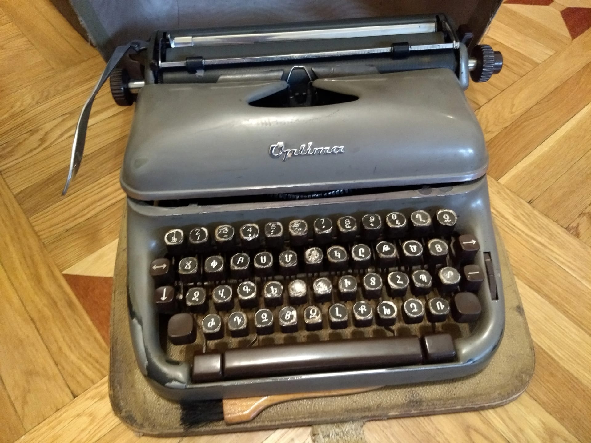 Eine graue mechanische Schreibmaschine mit armenischer Tastatur, in ihrem Lederkoffer auf einem Holzboden.